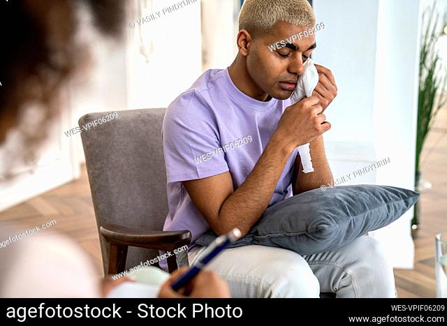 Sad patient sitting in armchair wiping tears with facial tissue