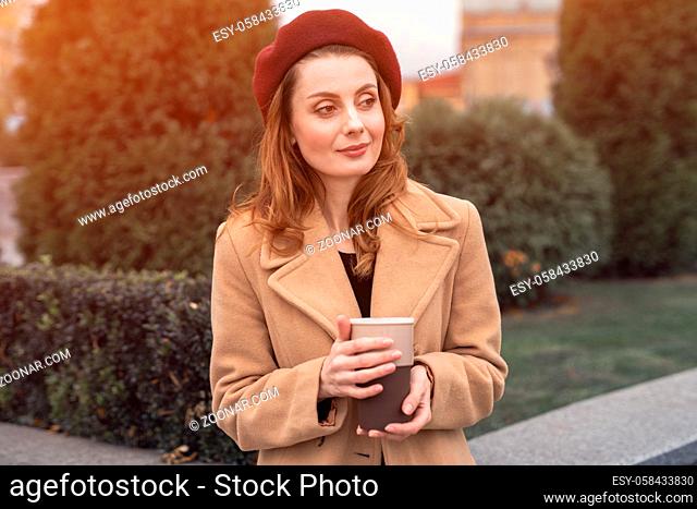 Pensive beautiful young woman holding a cup of coffee on the street. Female fashion concept. Portrait of stylish young woman wearing autumn coat and red beret...