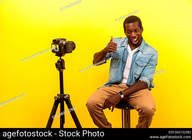 Put like for live blog. Portrait of happy friendly man in denim clothes smiling at his professional dslr camera and showing thumbs up, filming video
