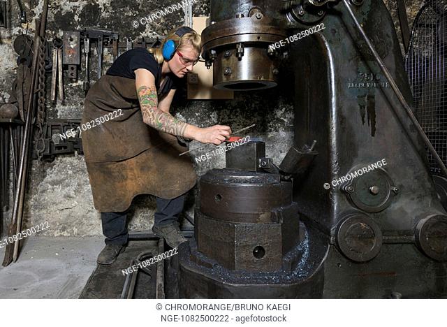female blacksmith measuring a glowing piece of steel, steam hammer