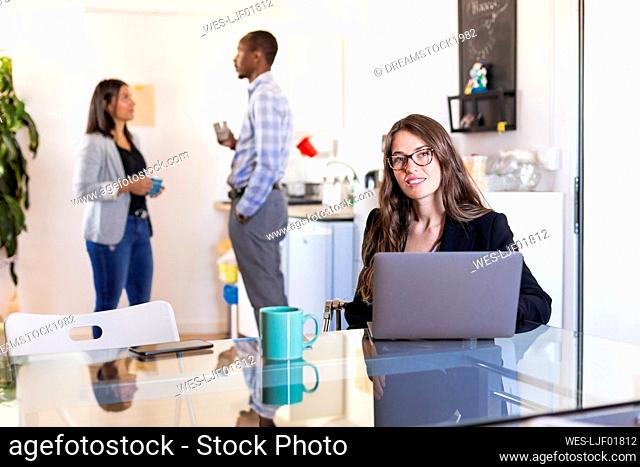Smiling businesswoman sitting with laptop at table while colleagues discussing in background during coffee break