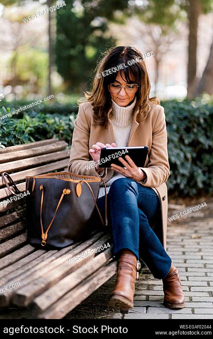 Mature woman using digital tablet while sitting on bench at park