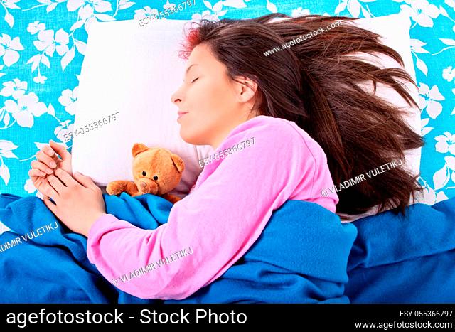 Close up of a beautiful young girl in pink pajamas sleeping in her bed, hugging a cute plush teddy bear. Sleep and relaxation concept