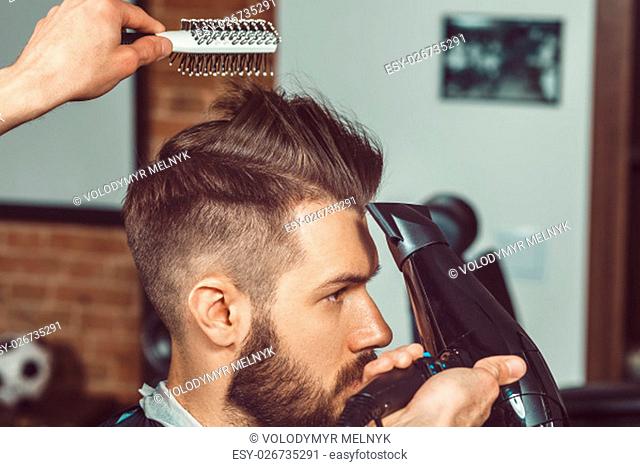 The hands of young barber making haircut of attractive bearded man in barbershop