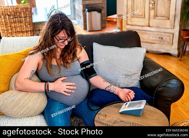 A high angle shot of a pregnant woman in her thirties, checking blood pressure with a digital device as she feels the baby kicking with hand on belly