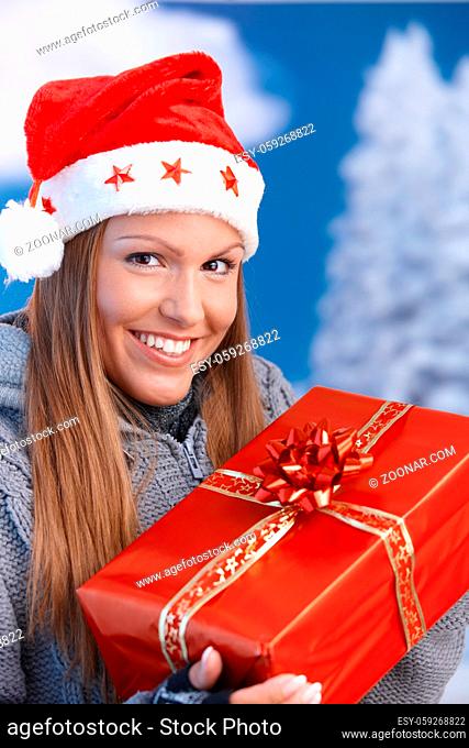 Happy young woman in santa hat holding christmas present, looking at camera, smiling