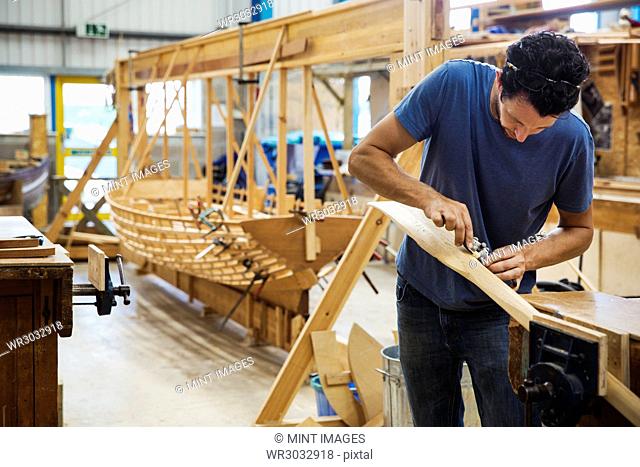 Man standing in a boat-builder's workshop, working on a wooden oar