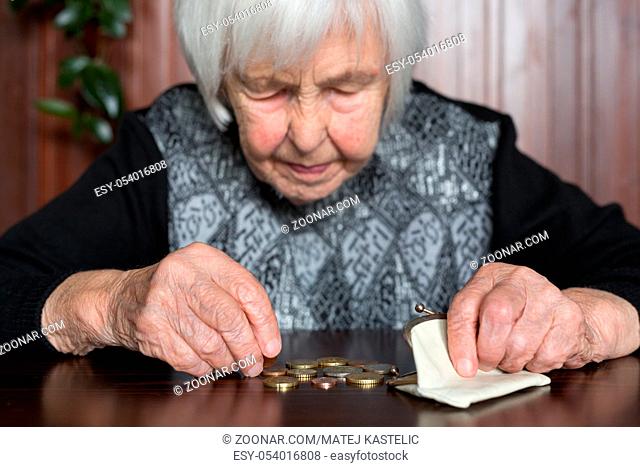 Elderly 95 years old woman sitting miserably at the table at home and counting remaining coins from the pension in her wallet after paying the bills