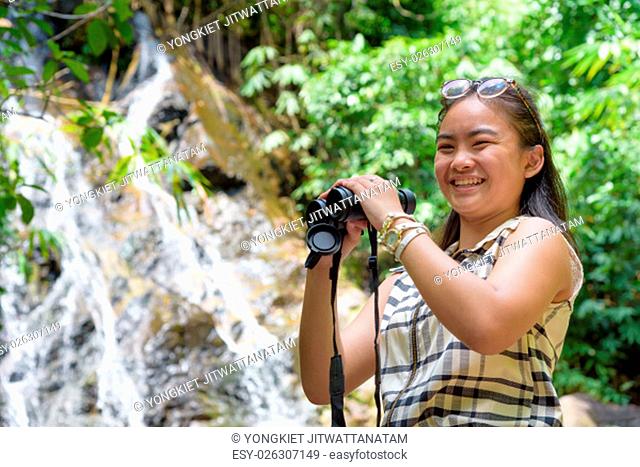 Beautiful young girl hiking is using binoculars look for birds smiling to the camera in tropical forest near the waterfalls in Thailand