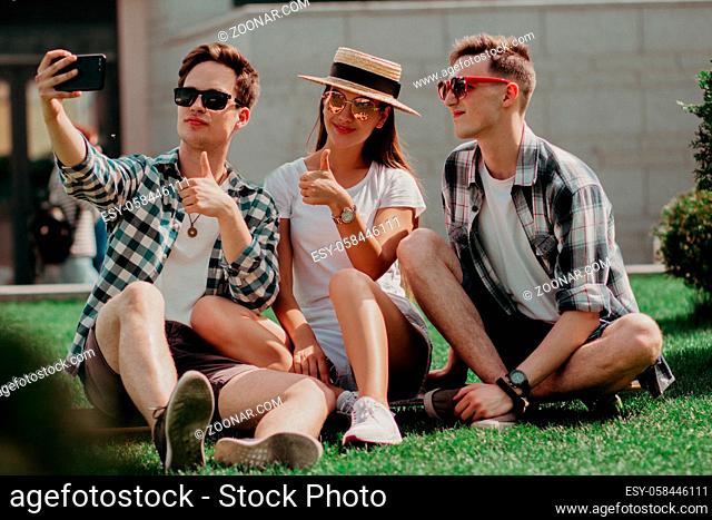 Three Light-Skinned Guys Pose For Selfies Sitting On The Lawn On A Summer Day. Two Attractive Boys And A Girl In A Hat And Sunglasses Take Pictures And Relax On...