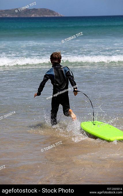Carefree boy in wet suit running with body board in sunny ocean water