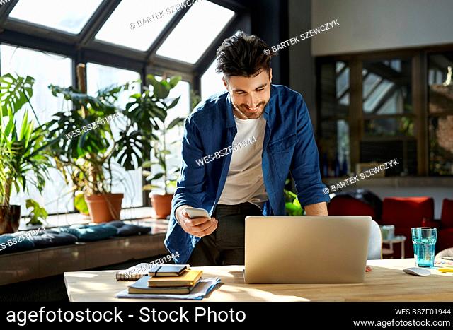 Smiling businessman with smart phone talking during video conference through laptop in office