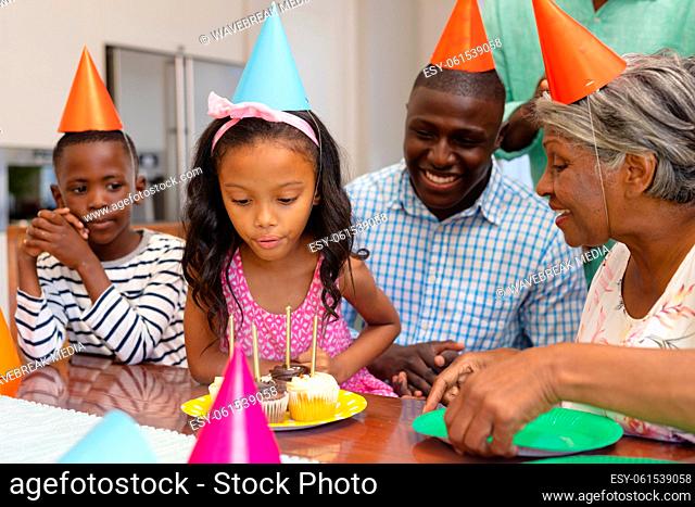 Multiracial excited girl wearing hat blowing candles while celebrating birthday with family at home