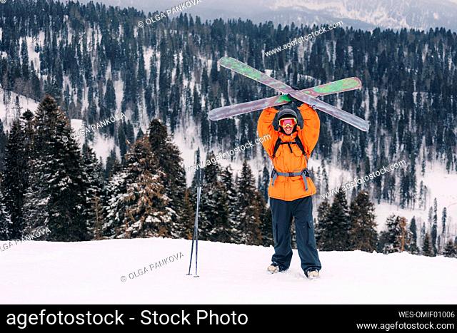 Smiling skier holding skis standing with arms raised in front of mountains