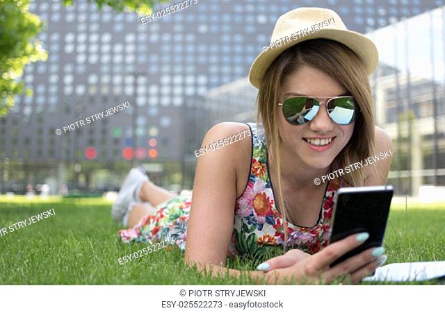 Stylish Attractive Girl Wearing Hat and Sunglasses, Using her Mobile Phone While Lying on the Grasses with Happy Facial Expression