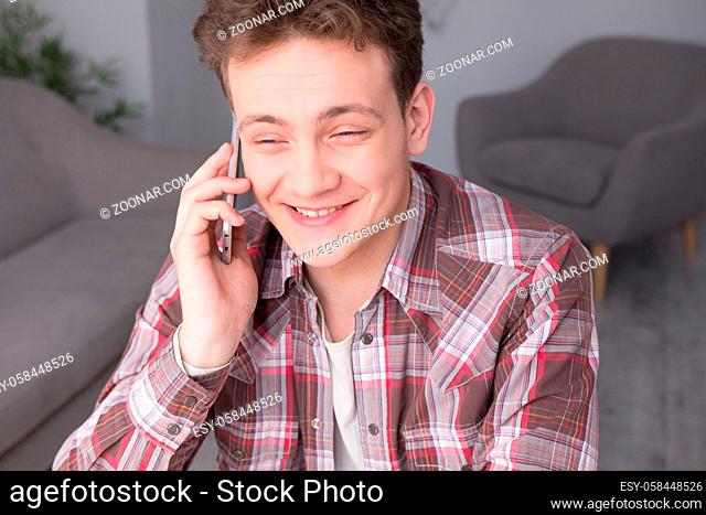 Young guy talking on phone and laughing. Teenager wearing long sleeved button up shirt smiling while making phone call indoors