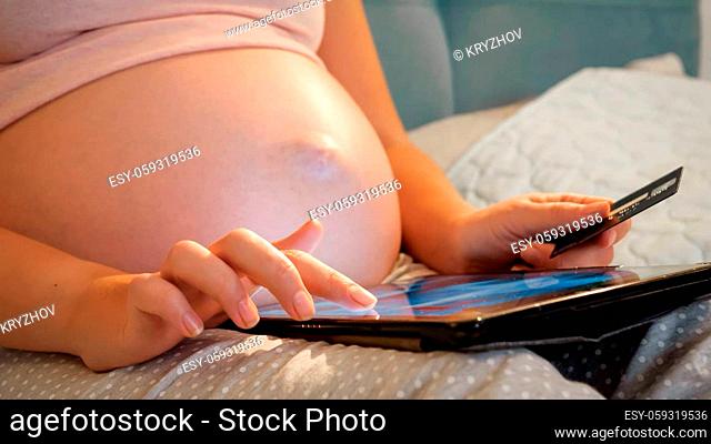 Closeup of pregnant woman browsing internet on tablet holding credit card. Woman expecting baby doing online shopping and purchasing clothes online