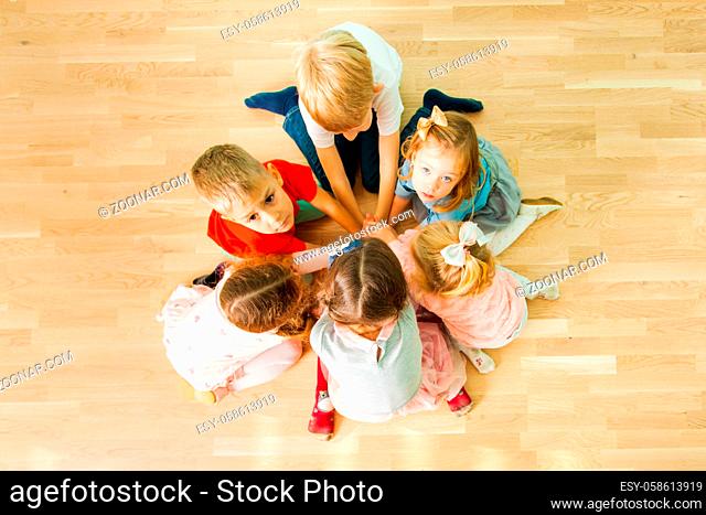 Top view of six kids sitting closely in a circle playing at the kindergarten. Group of children kneeling on a floor with their hands put together