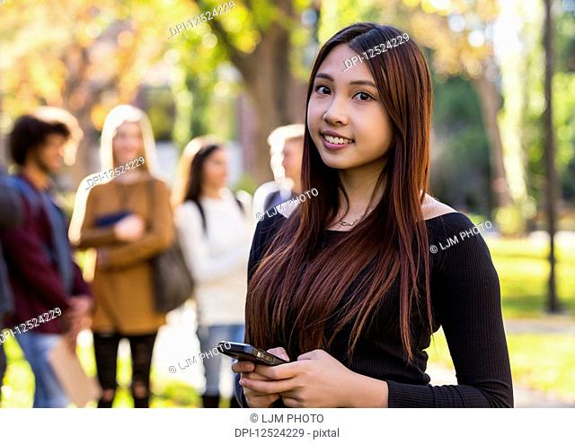 Portrait of a young female International university student holding a smart phone with her friends standing in the background; Edmonton, Alberta, Canada