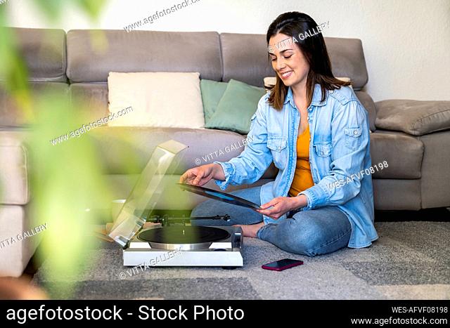 Smiling woman with turntable holding record while sitting by sofa at home