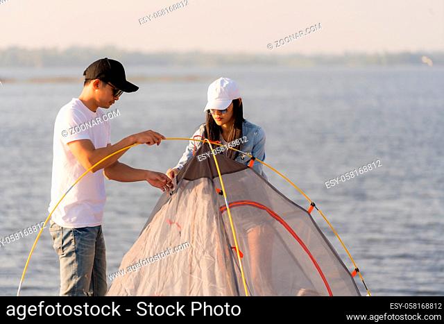 Young adult Asian couple pitch and set up tent for camping around the lake. Camping trip with dog outdoor activity campsite concept