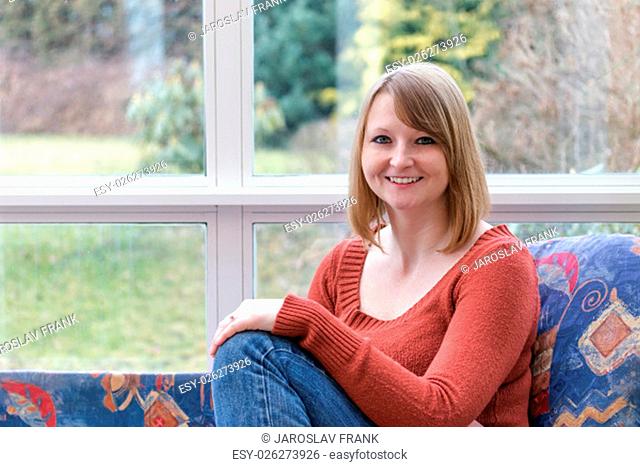 Smiling young woman dressed in a brown sweater is sitting on the sofa in conservatory and she is looking at the camera