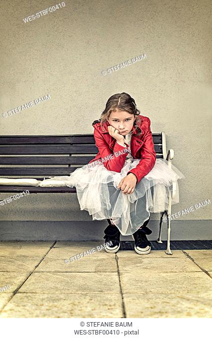 Portrait of unhappy girl wearing red leather jacket and tutu sitting on bench