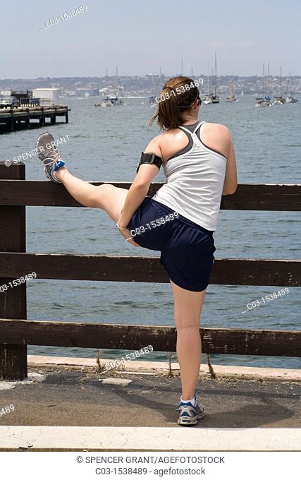 A woman jogger stretches her hamstrings on a fence in San Diego, CA