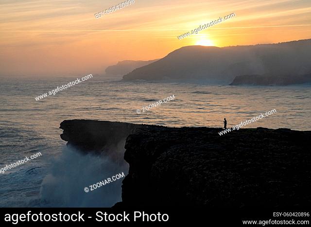 A view of huge storm surge ocean waves crashing onto shore and cliffs at sunrise with a person standing there