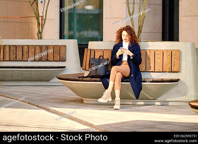 Serious woman sitting on bench and checking message on her smartphone outdoors