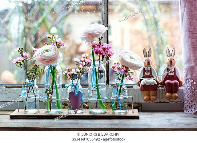 Turban Buttercup (Ranunculus asiaticus) and Geraldton Wax (Chamelaucium uncinatum). Small bottles with flowers, heart and decorative Easter bunnies in a window