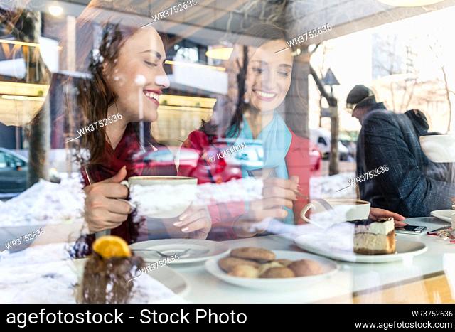 Two young and beautiful women smiling while enjoying together delicious cakes and coffee in a trendy cafe downtown