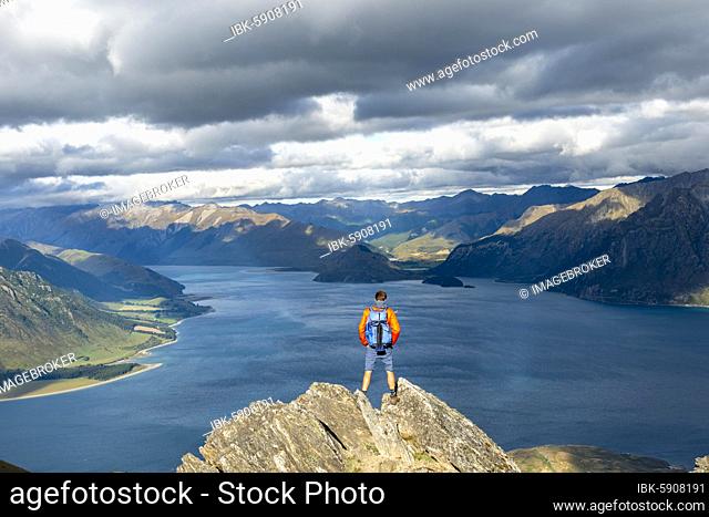 Hiker stands on a rock, view over Lake Hawea, lake and mountain landscape in the evening light, view from Isthmus Peak, Wanaka, Otago, South Island, New Zealand