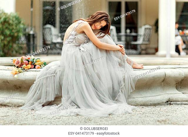 Pretty bride with long curly hair in wedding dress sitting near fountain at park background, wedding photo, portrait, close up