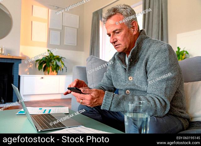 Focused caucasian senior man sitting on sofa, doing paperwork, using smartphone and laptop