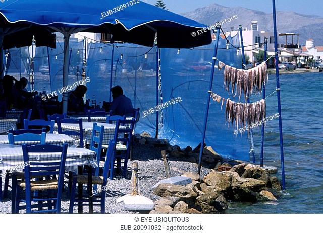 Restaurant outside seating with blue table and chairs next to waters edge with a fresh catch of seafood hanging from poles
