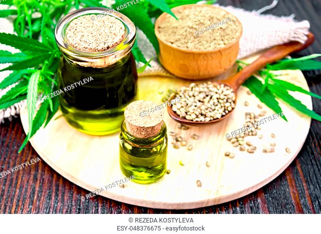 Hemp oil in two glass jars, grain in a spoon, flour in a bowl, leaves and stalks of cannabis, a napkin of burlap on a wooden board background