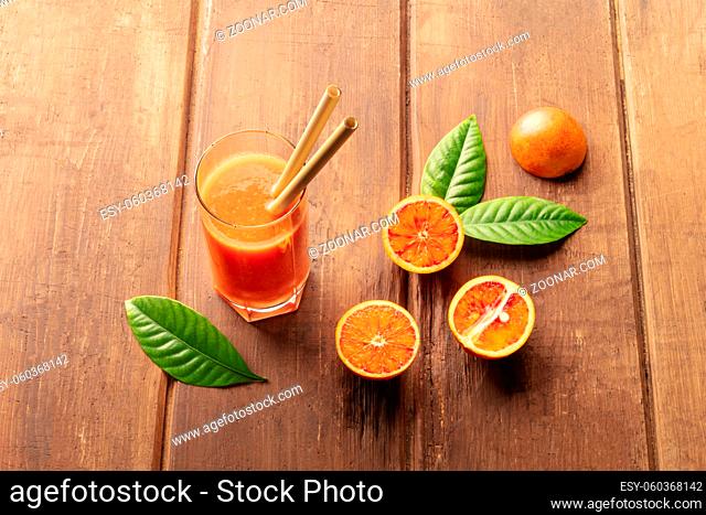A glass of fresh orange juice with blood oranges, green leaves, and two bamboo straws, on a dark wooden background with a place for text