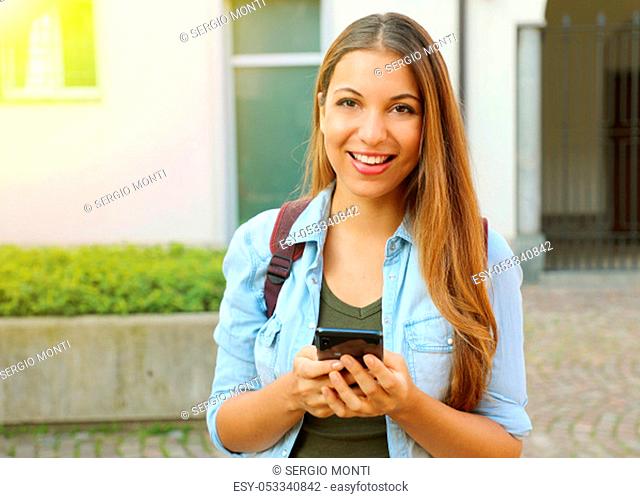 Beautiful young student girl typing on smart phone and looks at camera
