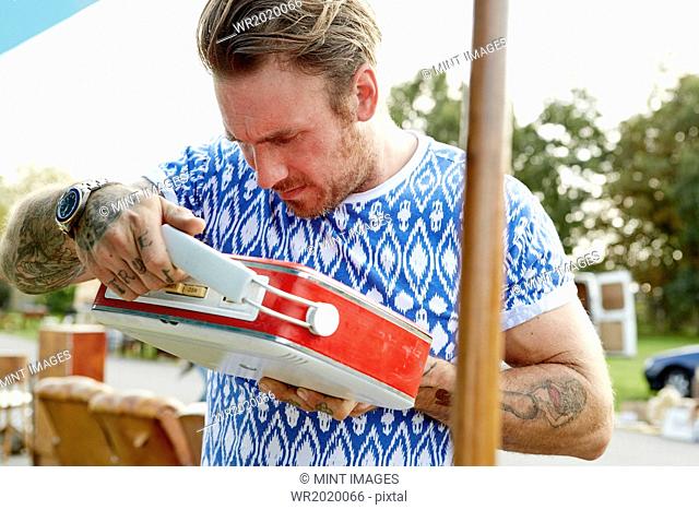 A man in a blue shirt with tattooed forearms, looking at a red vintage radio at a flea market