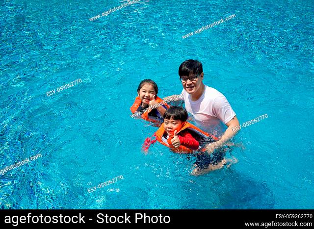 Asian father teaches his son and daughter to swim in the pool. The older sister and younger brother wearing orange life jacket, smiling with thumbs up