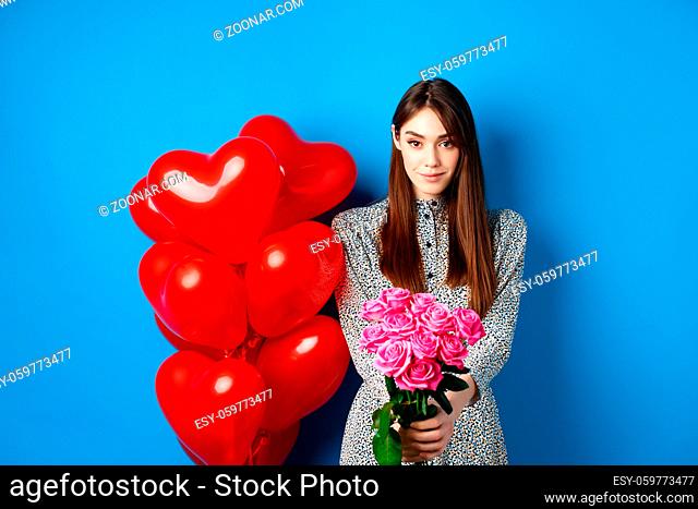 Valentines day. Pretty woman making romantic gift to girlfriend, stretch out hands with beautiful pink flowers and smiling, standing on blue background