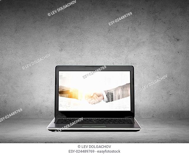 cooperation, partnership, technology and business concept - laptop computer with handshake on screen over gray concrete background with double exposure effect