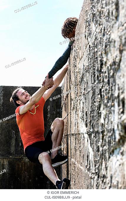 Young male free climber at top of sea wall helping friend climb up