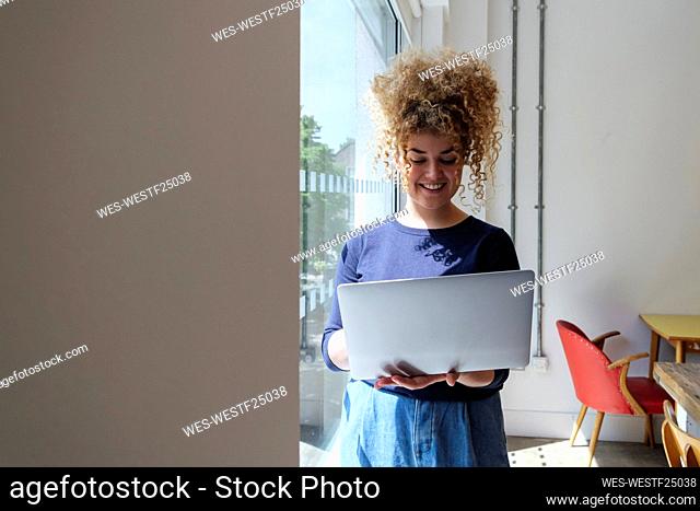 Young businesswoman standing by window holding laptop