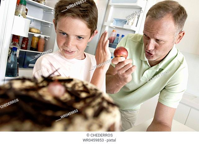 Boy standing in front of a cake and refusing an apple from his father