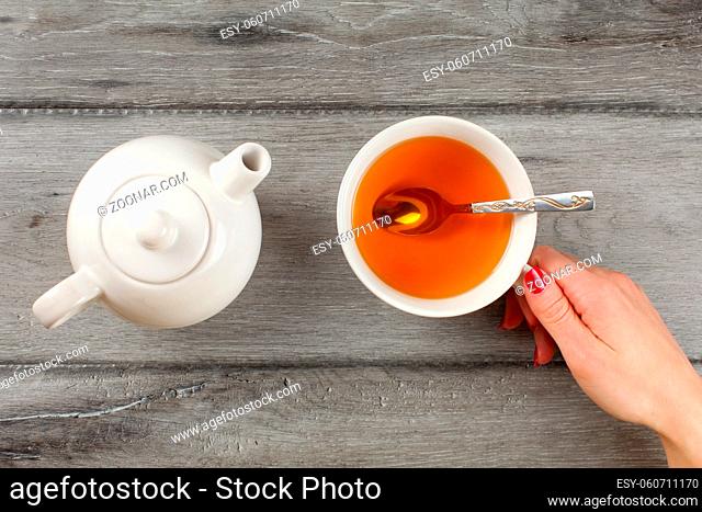 Top view, young woman hand with red nail holding cup of amber tea, with teapot next to it