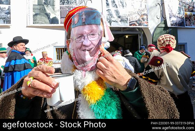 01 March 2022, Baden-Wuerttemberg, Riedlingen: A jester wearing a paper mask showing Minister President Winfried Kretschmann (Greens) stands outside with a cup...