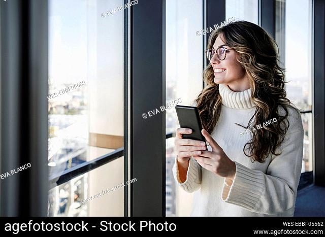 Happy businesswoman holding smart phone near window in office