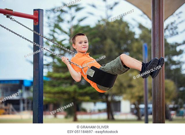 A side-view shot of a young caucasian boy wearing casual clothing, he is having fun on a swing in a public park
